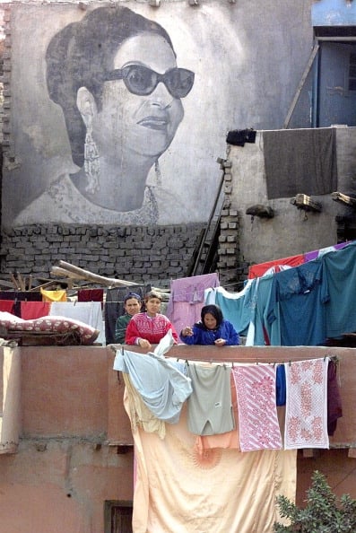 Egyptian women collects clothes in front of a giant picture of Egyptian diva Umm Kulthum painted on a shack in the Kom al-Ghurab suburb of Cairo 02 February 2000. Egypt is marking the 25th anniversary of Oum Kalthoum's death with a series of ceremonies to honor the star whose two songs Inta Omri and Al-Atlal still top the charts of bestselling Arab songs across the Arab world. Both songs were released in the sixties. (ELECTRONIC IMAGE) (Photo credit should read MARWAN NAAMANI/AFP/Getty Images)